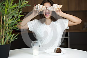 Funny young woman holding two croissants near her eyes showing tongue while having breakfast in the morning