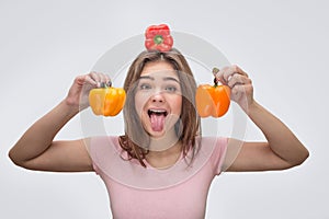 Funny young woman look on camer and show tongue. She hold red peeper on head and two yellow ones on each side of head