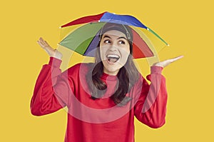 Funny young woman is protected from rain or sun with help of rainbow umbrella hat on her head.