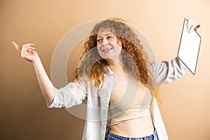 Funny young woman with red curly long hair holding hands aside and in one hand kepping tablet with white screen over photo