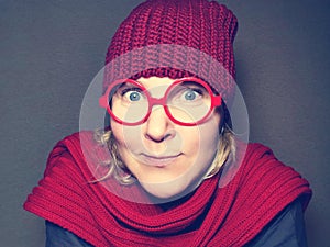 Funny young woman in red round glasses, red knitted hat and scarf grimaces looking at the camera. Comical image.