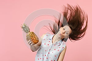 Funny young woman in summer clothes with flowing hair holding fresh ripe pineapple fruit isolated on pink pastel wall