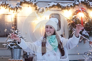 Funny Young Woman in winter forest while holdes a sparkler in her hand. Wearing stylish white coat and warm hat. Festive mood