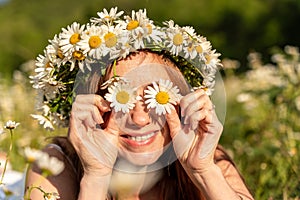A funny young woman in a wreath of daisies is laughing and holding daisies in front of her eyes. On a large field of daisies