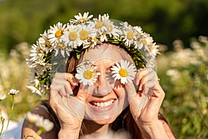 A funny young woman in a wreath of daisies is laughing and holding daisies in front of her eyes. On a large field of daisies