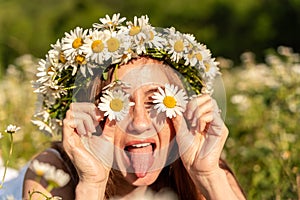 A funny young woman in a wreath of daisies is laughing and holding daisies in front of her eyes with her tongue hanging out. On a