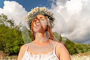 A funny young woman in a wreath of daisies is laughing and holding daisies in front of her eyes with her tongue hanging