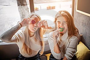 Funny young women cover eyes with macarons. They pose on camera. Brunette bites piece of macarone. They have fun.