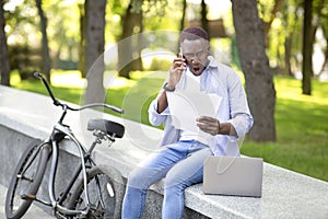 Furious African American guy with papers and laptop screaming into smartphone near his bike at park