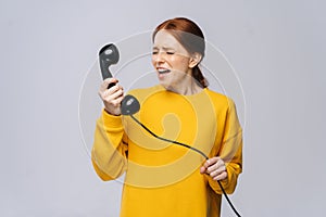 Furious angry young woman in stylish yellow sweater talking on retro phone and screaming in handset.