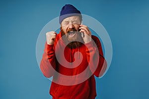 Furious man shouting and clenching his fist while talking on mobile phone isolated over blue wall