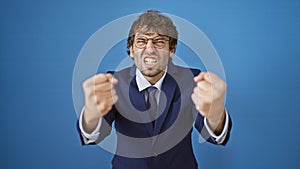 Furious, frustrated young man in business suit, fists raised in rage, screaming his anger, against blue isolated background