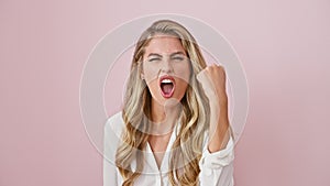 Furious young blonde, fists clenched in rage, agitatedly screams over isolated pink background. madly angry woman standing, shirt-