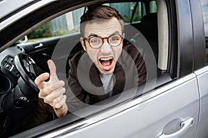 Furious young man is looking at the camera while sitting at his car. He is screaming at someone. Angry driver concept