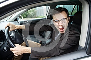 Furious young man is looking at the camera while sitting at his car. He is screaming at someone. His hands are on the steering