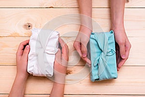 Furoshiki eco-friendly gifts wrapped in cloth in children`s hands on a wooden background. Zero waste. Top view