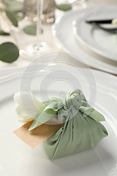 Furoshiki technique. Gift packed in green fabric with flower and blank card on table, closeup
