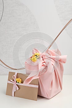 Furoshiki technique. Gift packed in pink fabric, card, and flowers on white table