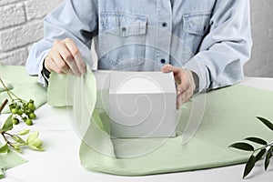 Furoshiki technique. Woman wrapping gift in green fabric at white table, closeup