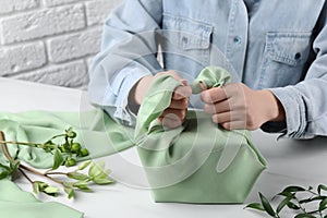 Furoshiki technique. Woman wrapping gift in green fabric at white table, closeup