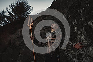 A fury man with a trident in his hands against the background of rocks.