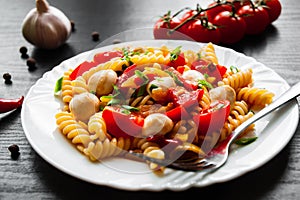 Pasta salad with tomato, pepper and mozzarella balls in plate on dark wooden background