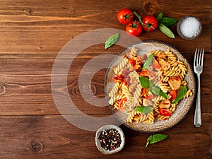 Fusilli pasta with tomato sauce, chicken fillet with basil leaves on dark brown wooden background, copy space, top view