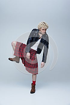 Fusion of cultures. Full length portrait of man in Scottish kilt and punk leather jacket posing against grey studio