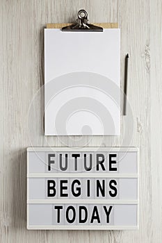 `Future begins today` words on a modern board, clipboard with blank sheet of paper on a white wooden surface, top view. Overhead