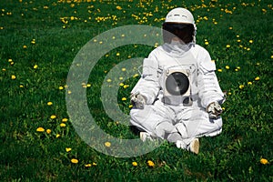 Futuristic astronaut in a helmet sits on a green lawn among flowersin a meditative position