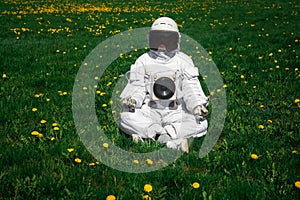 Futuristic astronaut in a helmet sits on a green lawn among flowersin a meditative position