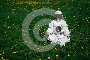 Futuristic astronaut in a helmet sits on a green lawn among flowersin a meditative position