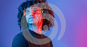 In futuristic glasses. Young beautiful man with curly hair is indoors in the studio with neon lighting