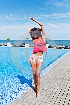 Fuul-lengh photo of attractive brunette girl with long hair posing to the camera near pool. She wears pink T-shirt with