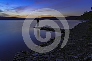 Fuzzy unfocused silhouette of man in fishing process on pier coast line lake waterfront in twilight lighting after sunset time