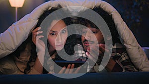 Gadget addict multiracial couple diverse man and woman scrolling mobile phone at night under blanket confused talking