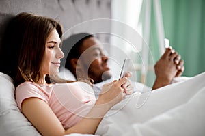Gadget addiction concept. Young multiethnic family using smartphones while lying in bed, copy space