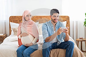 Gadget addiction concept. Young muslim man playing on smartphone and neglecting his expectant wife, sitting on bed