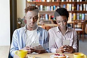 Gadget addiction and phubbing. Young black couple having dull date, stuck in smartphones, ignoring each other at cafe photo