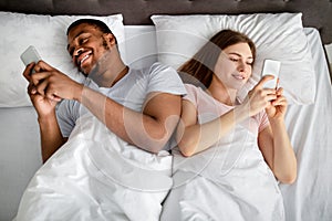 Gadget addiction and relationship problems. Millennial interracial couple with smartphones lying in bed, top view