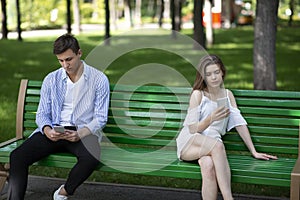 Gadget addiction and relationship problems. Young couple with smartphones sitting on bench at park, ignoring each other