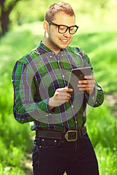 Gadget freak concept. Portrait of a funny hipster in casual clothing and trendy glasses reading e-book in the park. White shiny