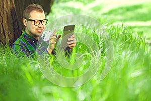 Gadget freak concept. Portrait of funny hipster in casual clothing and trendy glasses reading e-book sitting in green grass under