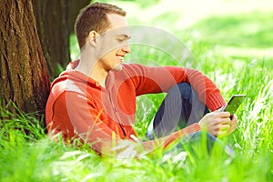Gadget freak concept. Portrait of smiling young man in casual clothing reading e-book, sitting in green grass under tree in the