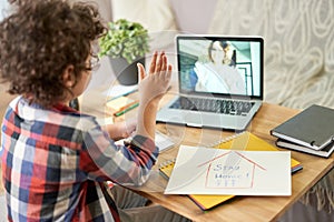 Gain the knowledge you seek. Rear view of hispanic school boy waving hand at teacher during online lesson via video chat