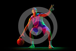 Game strategy. Young guy, basketball player in uniform dribbling ball against black studio background in neon light
