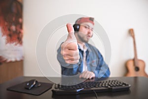 Gamer in the cap and headphones sits at home on the computer and shows the thumb up. Focus on your finger photo