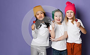 Gang of three cool kids in white t-shirts, colorful hats and pants posing with lollipops hugging together on purple