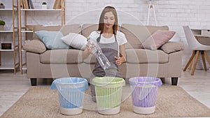 Garbage recycling concept. Young caring woman putting plastic bottle into green dustbin and smiling to camera at home