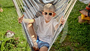 In a garden, a delighted boy wearing sunglasses enjoys the swing of a hammock, relishing the essence of summer, the